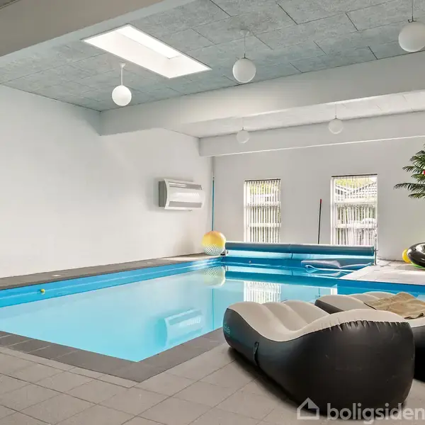 Indendørs swimmingpool med lænestole ved siden af. Omgivet af flisegulv og planter, med loftslamper og ovenlys. To store vinduer i baggrunden tillader naturligt lys.