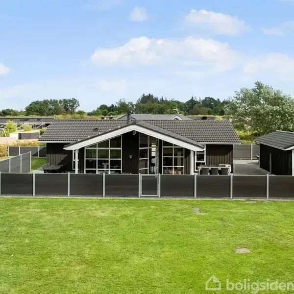 Sort træhus med store vinduer står stille på en indhegnet græsplæne. Til højre er et mindre skur. Baggrunden er en åben, grøn landskab med nogle træer og flere huse.