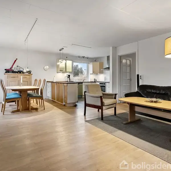 Et åbent stue- og køkkenområde viser en sort lædersofa og to borde. Et spisebord med stole står ved et vindue. Lyse farver dominerer rummets moderne design.