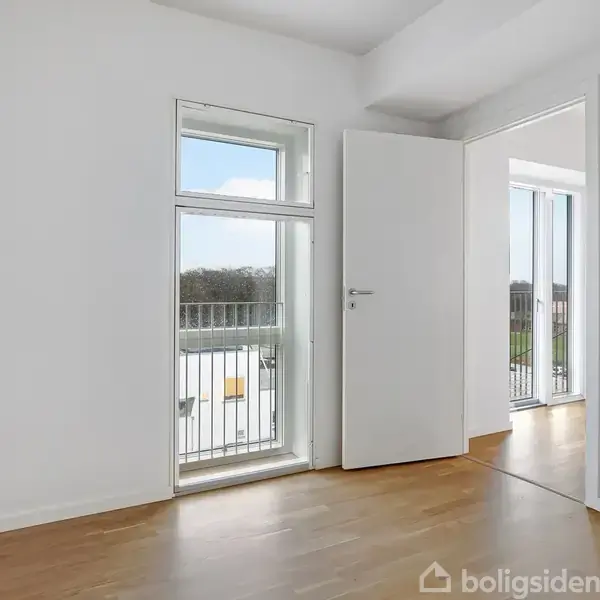 Et lyst værelse med trægulv, en åben dør og et stort vindue ud mod en balkon. Udenfor ses en del af landskabet.