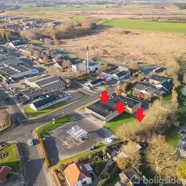 Tre røde pile peger på en række bygninger. Bygningerne er placeret i et beboelsesområde med veje og huse, omgivet af åbne marker og træer.