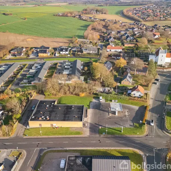 Bebyggelse set fra luften, med huse og en kirke spreder sig langs veje. Omkringliggende marker og træer omkranser området. Forreste bygninger ligger ved en hovedvej med synlig fortov.