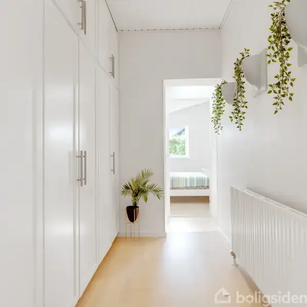 Gangen har hvide skabe og en radiator på væggen, med planter monteret over. En åben dør viser et værelse med seng og vindue i baggrunden.