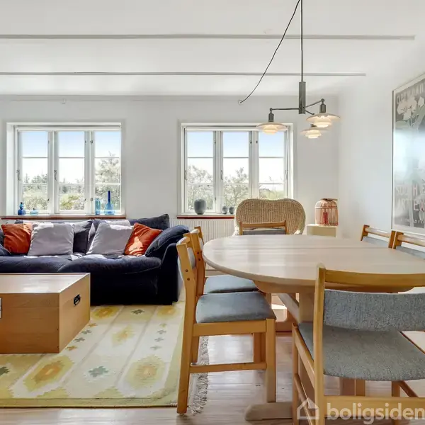 En stue med en blå sofa og sofabord på et mønstret tæppe. Ved siden af er et spisebord med stole, under en moderne lampe. Vinduerne i baggrunden giver naturligt lys.