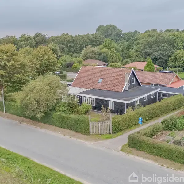 Et sort hus med rødt tag står stille ved en vej. Omkring huset er der grønne hække og træer i en landlig, skovklædt område.