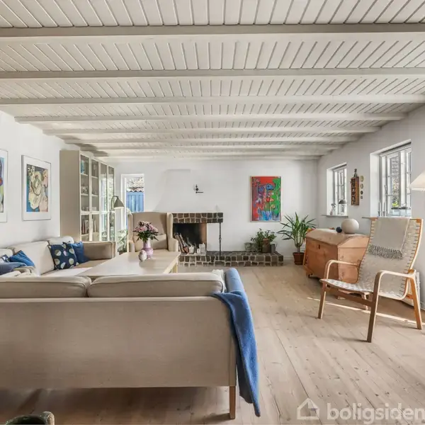 Stue med cremefarvet sofa og blå tæppe foran pejsen. Der er kunst på væggene, træmøbler og planter i lys, åben atmosfære med hvide trælofter og store vinduer.