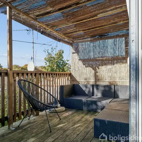 Terrasse med sort kurvestol og grå sofa placeret under en pergola med bambusloft, set mod grønne træer i baggrunden. En hvid lanterne hænger fra loftet.