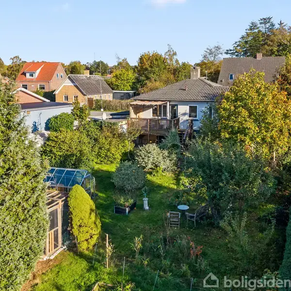 Et hus med en stor have omgivet af grønne træer og buske. Der er en terrasse mod baghaven, og forskellige små strukturer ses blandt tæt bevoksning i et boligområde.