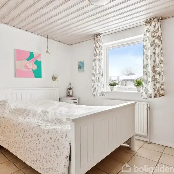 Seng står i et lyst soveværelse med hvidmalede vægge og loft. To billeder hænger på væggen over sengen, med et natbord ved siden af. Et vindue har udsigt udenfor.