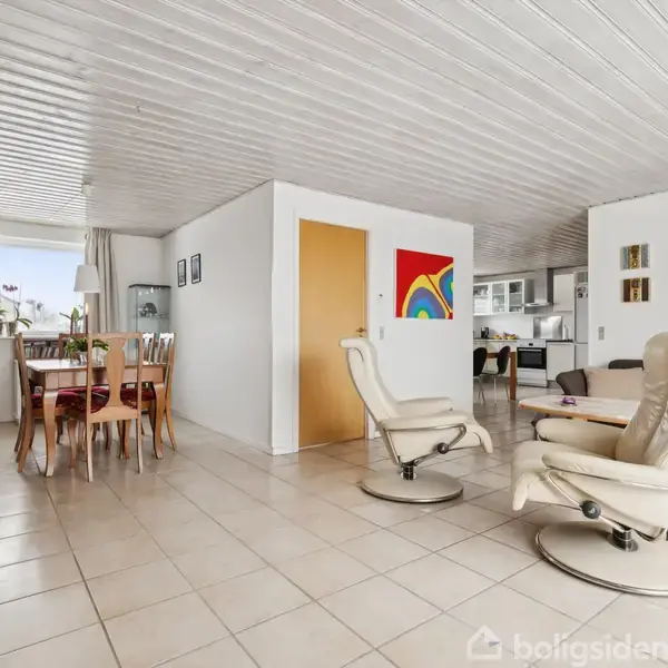 To lænestole står i et flisebelagt stueområde, hvor en farverig plakat hænger på væggen. I baggrunden ses spisebord med stole ved vindue.