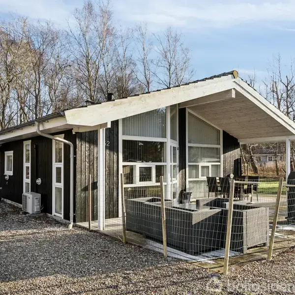 Et sort træhus med store vinduer har en terrasse med havemøbler, omgivet af et grusområde. I baggrunden ses bladløse træer og en klar blå himmel.