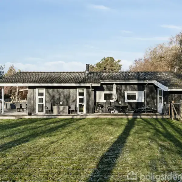 En sort træhytte står stille på en velplejet græsplæne. Foran hytten er der havemøbler, mens træer afslutter scenen om baggrunden.