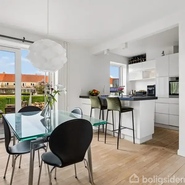 Spisebord med sorte stole står på trægulv ved siden af et åbent køkken med barstole. Store vinduer giver udsigt til en terrasse og have.