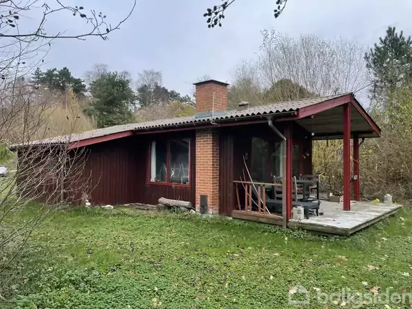 Et lille, træbeklædt sommerhus med en murstensskorsten står i en frodig have. Det har en veranda med tæpper og et overdækket område. Omkringliggende træer skaber en rolig atmosfære.