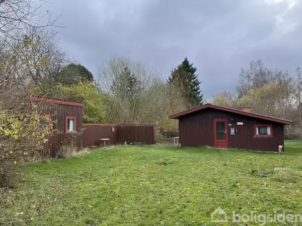 To træhytter står i en have med græsplæne og omgivende skov. Den ene hytte har et stort vindue; begge er malet mørkerød. Himmel er overskyet. Ingen tekst til stede.