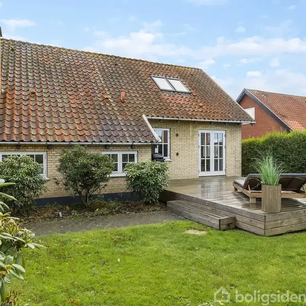 Blåt sofaarrangement står i en lys stue med store vinduer, trægulve og moderne spiraltrappe. En fladskærm hænger på en væg med træpaneler. Væggene pyntes af kunstværker.