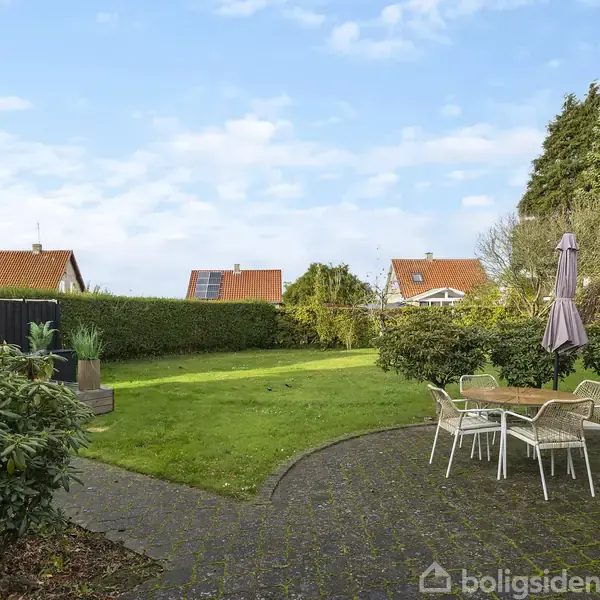 En have med en rund terrasse, hvor et bord og stole står under en parasol. Græsplænen omgivet af hække og buske. Murstenshuse med røde tage ses i baggrunden.