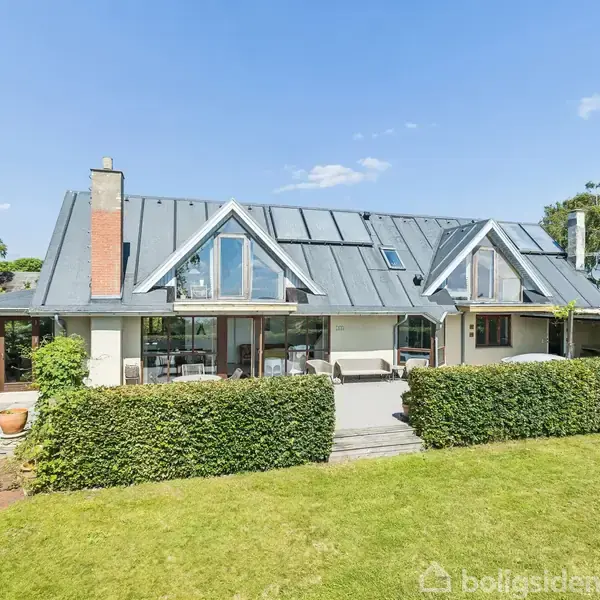 Et moderne hus med en metalbeklædt tag og store glaspartier står i en velplejet have med græs og buske. Himlen er klar og solrig.