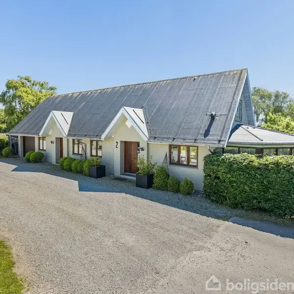 Et langstrakt hus med skrånende tag står i rolige omgivelser. Huset har hvid facade, flere vinduer, og en grusindkørsel foran. Omkranset af grønne buske og en mark i baggrunden. Husnummer 2 ses ved døren.