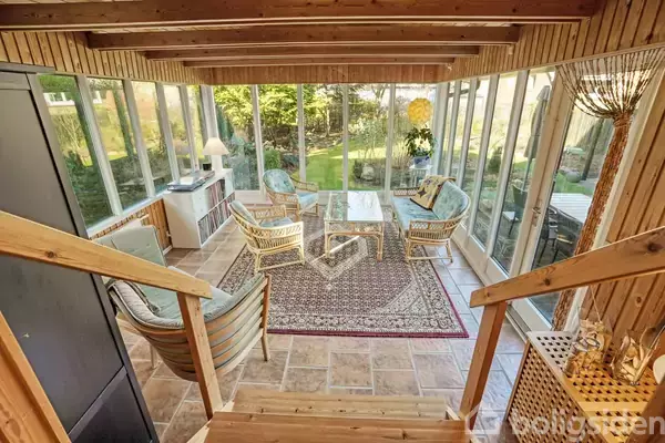 Træmøbler står på et tæppe i en glasindkapslet stue med udsigt til en grøn have. Et lille bordlampe og blomster dekorerer rummet, som har trægulv og trævægge.