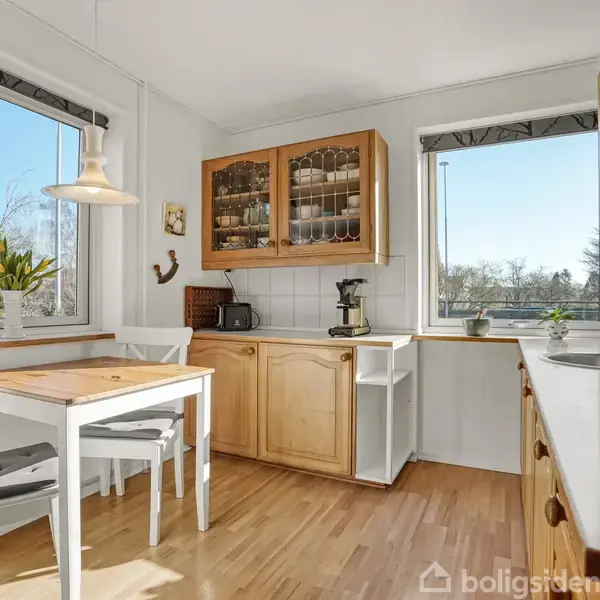 Et lyst køkken med træmøbler; et træbord og to stole står foran store vinduer. Et skab er monteret på væggen, og en vask er placeret på en lang bordplade. Sollys strømmer ind.