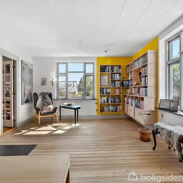 En stue har en brændeovn og et komfortabelt lænestol i hjørnet. En gul reol fyldt med bøger står op ad væggen. Store vinduer giver udsigt til en have.
