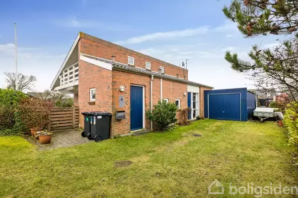 Stol står ved dør med glasparti, omgivet af planter i potter. Til venstre ses en åbning til et badeværelse med hvide møbler. Indrammet af murstensvægge og trægulv.