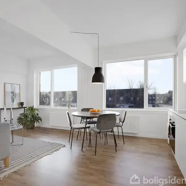 Et rundt bord med fire stole står i et lyst køkkenalrum. Store vinduer giver udsigt og naturligt lys. En sort lampe hænger over bordet, og planter pynter rummet.