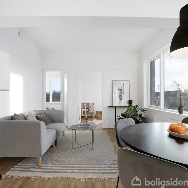 En moderne stue med grå sofa, sofabord og lampe. Et bord med appelsiner står i forgrunden. Store vinduer giver lys til rummet med minimalistisk indretning og planter.