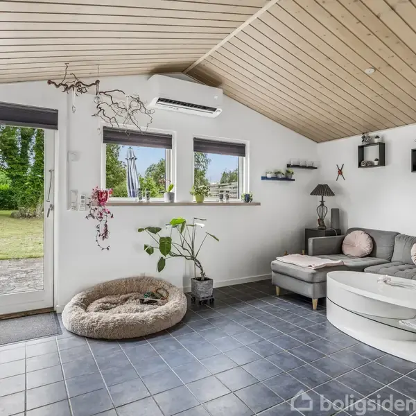 En stue med gråt klinkegulv og hvidmalede vægge. En grå sofa er placeret ved vinduet, med et rundt sofabord foran. Et stort vindue og en glasdør giver udsigt til en have.
