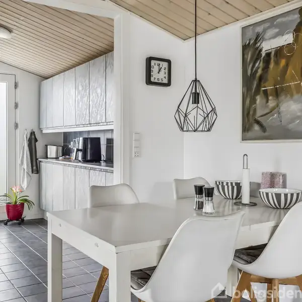Et spisebord med hvide stole står i et moderne køkken/alrum. En geometrisk lampe hænger over bordet. Klinkegulv strækker sig ind i et køkkenområde med lyse skabe og en plante.