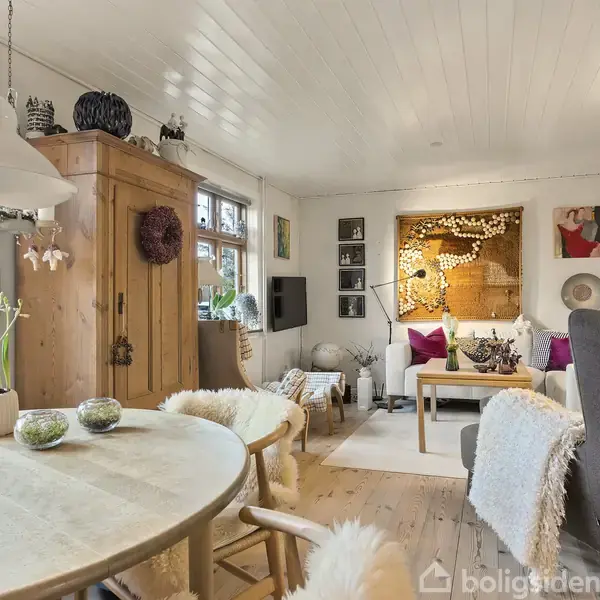 Spisebord med stole omkring det, dekoreret med planter og puder, står i en hyggelig stue med lyse træmøbler, kunst på væggene, og en sofa i baggrunden.