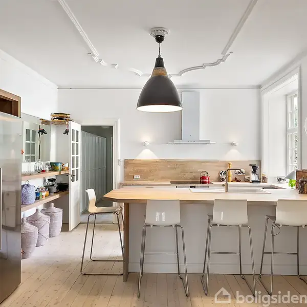Et lyst køkken med en central køkkenø omgivet af høje barstole. Der hænger en stor, sort lampe over bordet. I baggrunden ses skabe, hylder og hvide vægge.