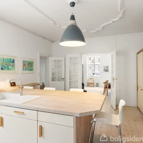 Et lyst køkken med en stor køkkenø med vask, omgivet af hvide skabe og en stålbordplade. En stor lampe hænger over øen. I baggrunden ses et spiseområde.