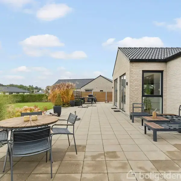 En moderne terrasse med flisebelægning har et rundt bord og stole til højre. I baggrunden ses græsplæne, planter og et hus i et villakvarter.