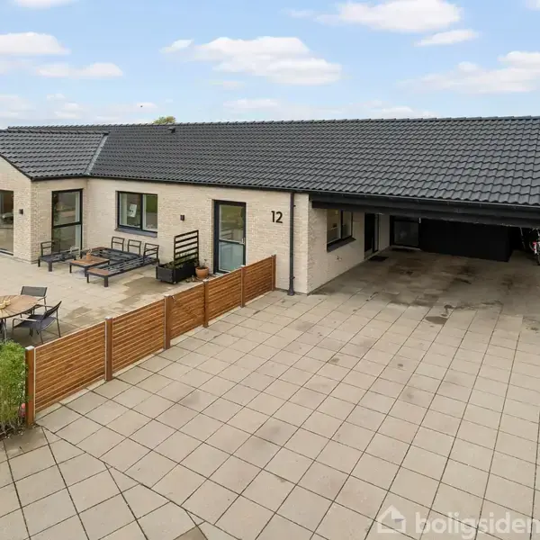 Hus med fladt tag, nummer 12, står i en flisebelagt gård med åbent garageområde til højre. Foran huset står havemøbler og et rundt bord, omgivet af lave hegn.