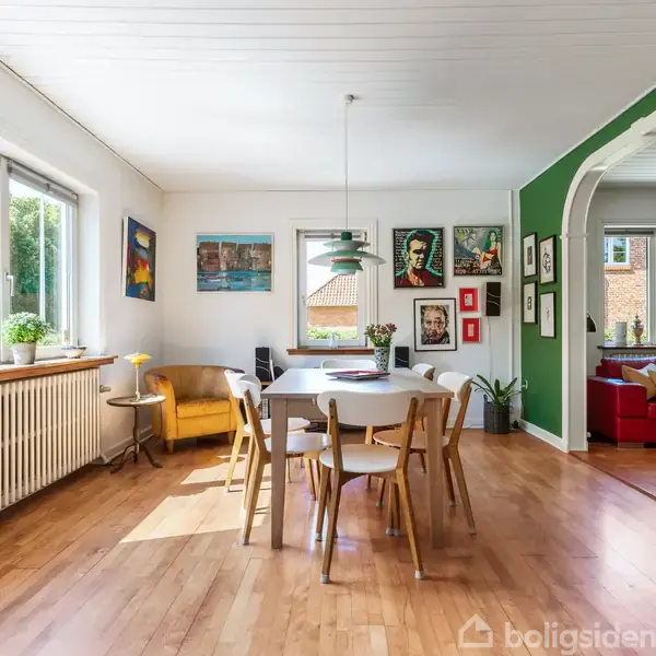 Spisebord med stole står centralt i et lyst rum med trægulve. Vægge dekoreret med kunst og grøn accentvæg. Store vinduer og adgang til opholdsstue med rød sofa.