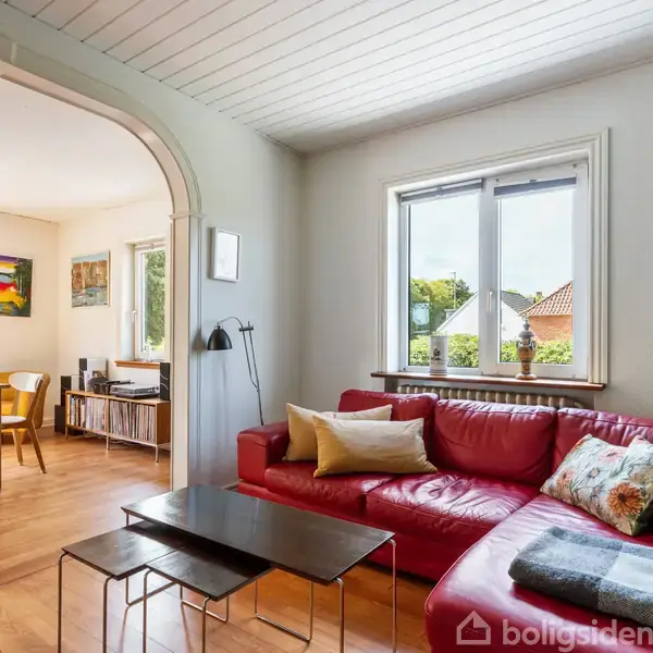 Rød sofa med puder står i en stue ved siden af et vindue, med udsigt til en have. Stuen fører ind til en spisestue med træstole og et bord.