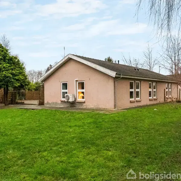 Et enkelt enfamiliehus med rosa facader har mange vinduer og ligger i en have med en stor græsplæne, omkranset af træer i et beboelsesområde.