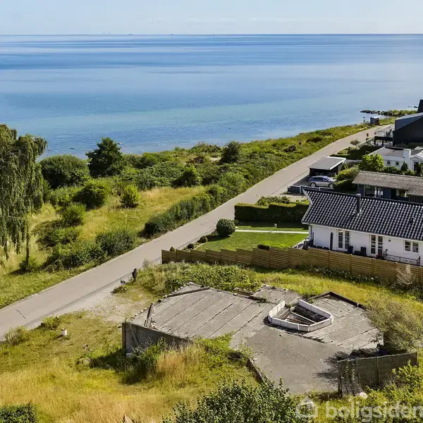 Cirkelformet betonfundament ligger ubenyttet ved en kystvej. Omkringliggende græsarealer og huse strækker sig mod en rolig blå havoverflade. Biler ses langs den snoede vej.