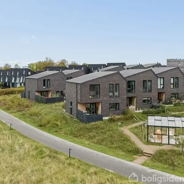 Moderne rækkehuse med sort facade står stille i et grønt boligområde. En asfalteret sti bugter sig gennem landskabet. En lille glasbygning er placeret i forgrunden.