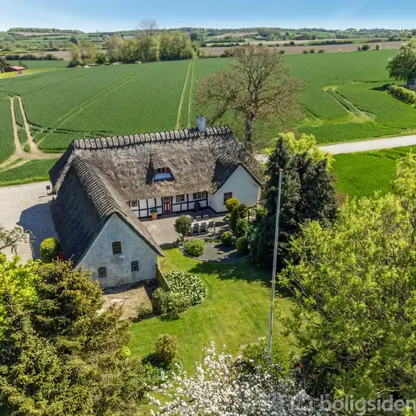 Bindingsværkshus med stråtag ligger omgivet af grønne marker og træer. Huset står på en velplejet græsplæne med buske og blomster, og en sti fører til en landevej i baggrunden.