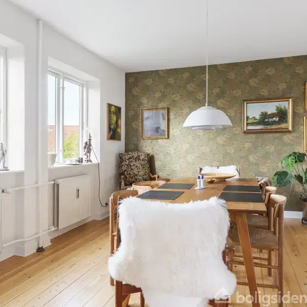 Spisebord med seks stole står i en lys stue med trægulve. Væggen har kunstværker, og der hænger en lampe over bordet. Store vinduer slipper naturligt lys ind.