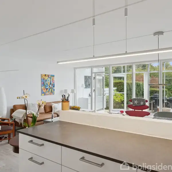 Køkkenbord står stille i lys stue med store vinduer og udsigt til grøn have. En sofa og lænestol giver komfort, mens moderne indretning skaber en ren og åben atmosfære.