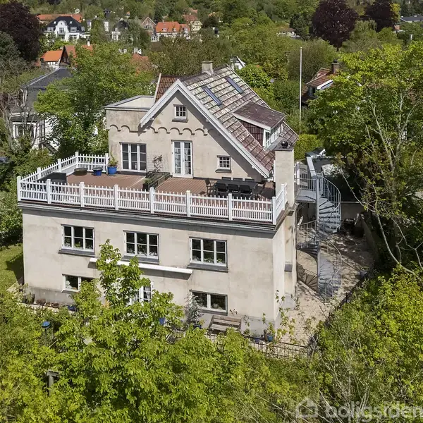 Et stort, hvidt hus med en tagterrasse omgivet af grønne træer i et villakvarter. Huset har flere vinduer og et skråtag med solpaneler.