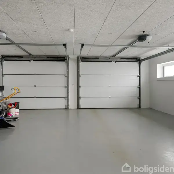 Et tomt garage med to lukkede, hvide porte. På venstre side står der legetøj og værktøj. Der er betongulv og et lille vindue til højre.