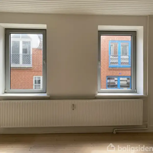 To vinduer i en hvid væg, med et varmeradiator nedenunder, ser ud på en bygning med røde mursten og blå vinduesrammer. Ingen tekst til stede.