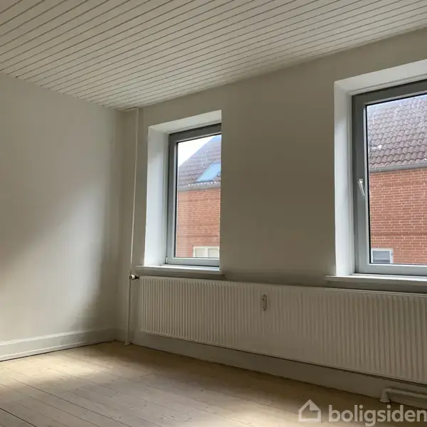 Et tomt værelse har to store vinduer med hvidrammede karme. En radiator er placeret under vinduerne. Udenfor ses røde murstensbygninger. Loftet er hvidt med træpaneler.