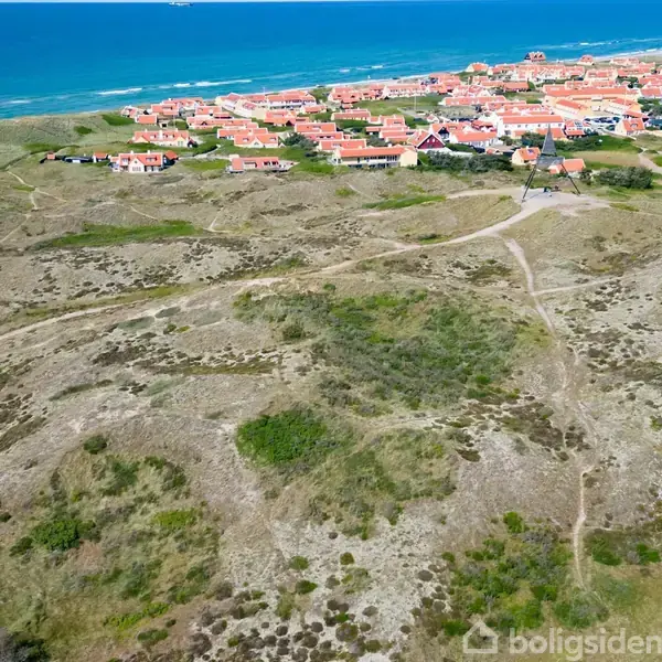 Kystby med røde hustage strækker sig langs klitterne ved havet. Stier snor sig gennem det sandede landskab mod blå bølger.