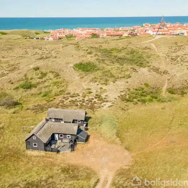 Et sort hus står stille i klitlandskab; omkring det er åbne sandklitter. I baggrunden ses en by med røde tage og havet. Ingen tekst bortset fra en udeladt vandmærke.